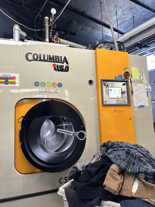 The industrial dry cleaning machine stands ready with its laundry basket brimming with clothes. Equipped with a detailed control panel, multiple labels, and visible pipes on top, this robust system ensures precise cleaning for every garment processed.